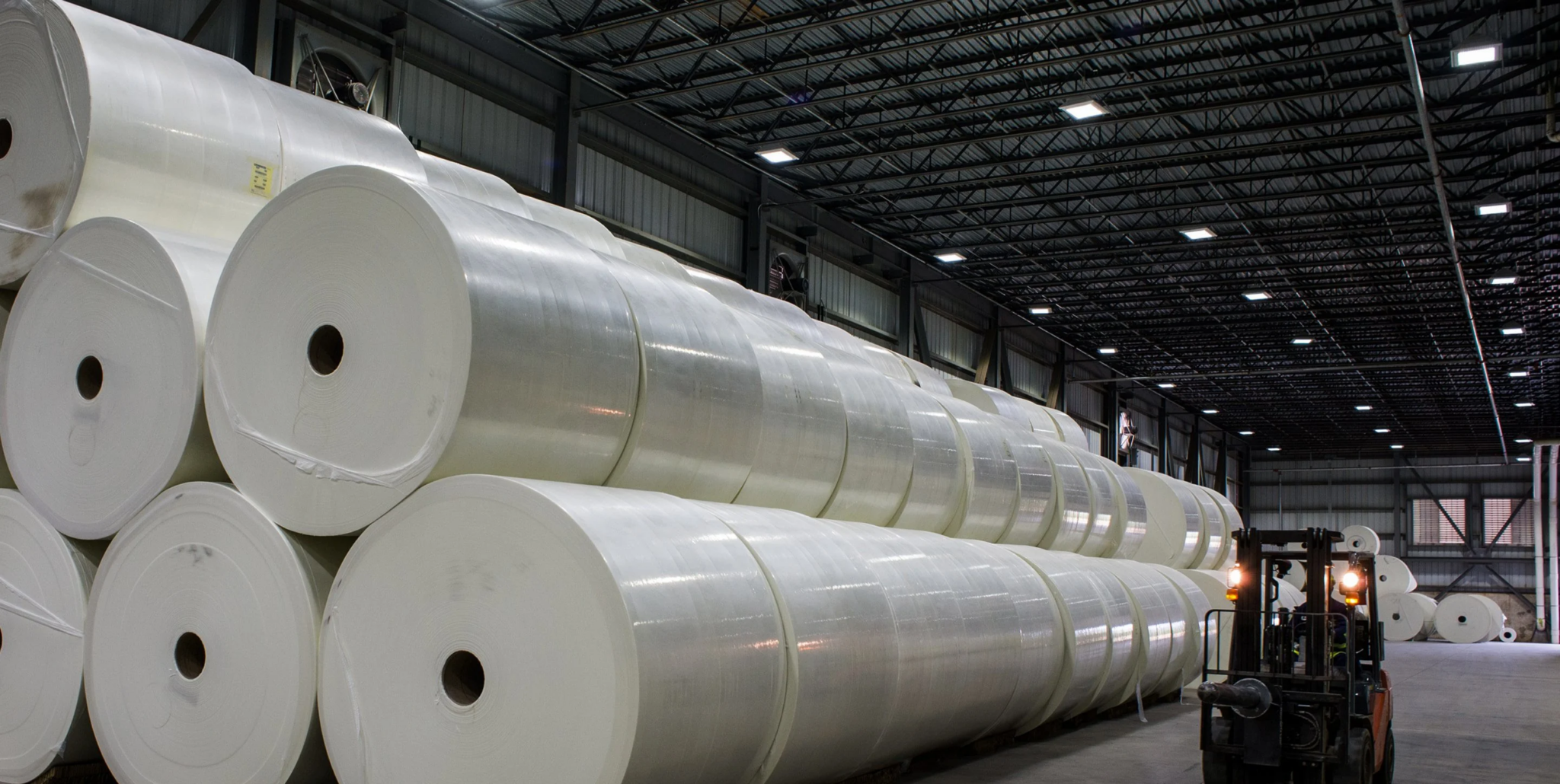 Large industrial warehouse interior stacked with giant white paper rolls and a forklift operating along the aisle
